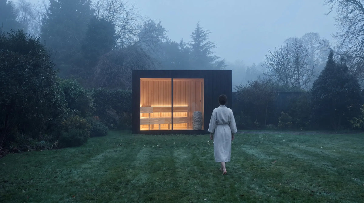 A lit NOOK Sauna in a misty garden, with someone in a robe walking towards it